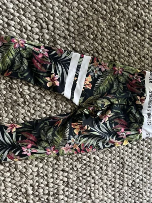 Blommiga tränings tights från STRONGER - Snygga leggings från Stronger med svart bas och tropiskt blommönster i grönt, rosa och gult. Breda vita ränder på ena benet och elastisk midja med logotyp. Perfekta för träning eller chill. Materialet är stretchigt och följsamt.