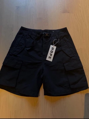 Svarta cargoshorts från HOPE, 35/44 (unisex) - Snygga svarta cargoshorts från HOPE med flera praktiska fickor och ett bälte i midjan. Shortsen har en loose passform och är perfekta för dig som gillar en avslappnad stil. Tillverkade i bomull för skön känsla. Unisex, Wom 36/Man44