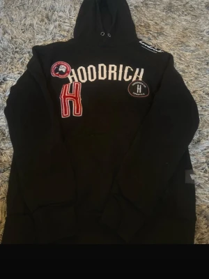 Svart hoodie från Hoodrich med patchar - Svart hoodie från Hoodrich med broderade patchar och tryck på bröstet. Stor röd bokstav H, rund patch och vit HOODRICH-text framtill. Klassisk huva med dragsko och ribbade muddar. Perfekt streetwear-stil för dig som gillar snygga detaljer. Snöre fattas dock.