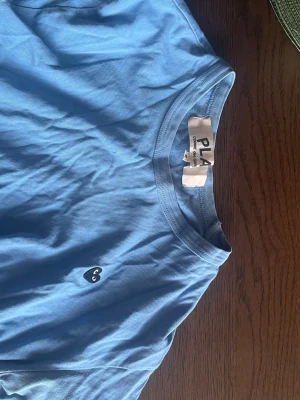 Blå t-shirt från Comme des Garçons Play - Snygg blå t-shirt från Comme des Garçons Play med det ikoniska hjärta-loggan broderad på bröstet. Klassisk passform med rund hals och korta ärmar. Perfekt för en stilren och avslappnad look.