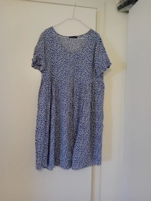 Blå blommig kortklänning - Supersöt blå kortklänning med vitt blommönster och rund halsringning. Klänningen har korta ärmar och ett luftigt, avslappnat fall. Perfekt för varma dagar och enkel att styla med sneakers eller sandaler.