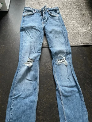 Mellanblå slitna jeans med slits - Säljer ett par mellanblå jeans med slitningar på båda knäna och råa, fransiga benslut från Zara. Jeansen har en liten slits vid benslutet och klassisk femficksmodell. Perfekta för en avslappnad och trendig look.