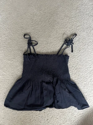 Mörkblå  linnetopp med knytband - Mörkblå svart linnetopp med smockad överdel och volang nertill. Axelbanden knyts på axlarna för en söt och justerbar look. Perfekt till jeans eller kjol för en trendig stil.