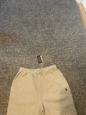 Gråa mjukisbyxor från Polo Ralph Lauren - Snygga grå mjukisbyxor från Polo Ralph Lauren med klassisk logga broderad på vänster ben. Byxorna har dragsko i midjan, två sidofickor och en bakficka. Perfekta för en chill och avslappnad stil. Tillverkade i mjukt och bekvämt material.
