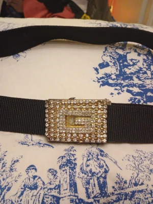 Svart bälte med glittrande spänne - Snyggt svart bälte med ett stort, fyrkantigt spänne täckt av glittrande stenar. Perfekt för att ge outfiten lite extra bling. Bältet har en enkel design men spännet gör det till en riktig statement-accessoar.