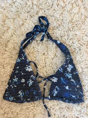 Blommig blå trekantsbikini - Supersnygg trekantsbikini med blå botten och vita blommor. Bikinin har smockad struktur, knyt i nacken och ryggen samt tunna band. Perfekt för strandhäng och sommarens alla bad.