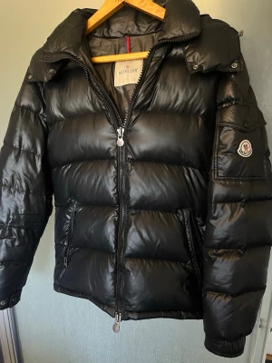 Svart dunjacka från Moncler - Snygg svart dunjacka från Moncler med glansig finish, hög krage och dragkedja framtill. Jackan har fickor med dragkedja och en ärmficka med Moncler-logga. Perfekt för kalla vinterdagar och riktigt varm tack vare dunfyllningen. Som du ser på näst sista bilden är där något smått hål därav priset.