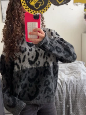 Grå leopardmönstrad stickad tröja - Mysig stickad tröja i grått med svart leopardmönster. Tröjan har en avslappnad passform, rund halsringning och långa ärmar. Perfekt för att hålla sig varm och snygg under kyliga dagar. Materialet är fluffigt och mjukt för extra komfort.