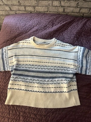 Vit och blå mönstrad stickad tröja - Mysig stickad tröja i vitt med blått mönster och ribbade muddar. Tröjan har rund halsringning och långa ärmar. Perfekt för kalla dagar och ger en avslappnad, trendig look.
