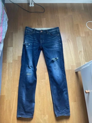 Mörkblå slitna jeans med raka ben - Snygga mörkblå jeans med raka ben och slitningar på låren och knäna. Klassisk femficksmodell med knapp och dragkedja. Jeansen har en cool tvätt och detaljerade slitningar för en trendig look. De är från Jack n Jones L32 W32 de ända flawn är hålet som är på bild där ser man lite hud men jag tycker de ba är snyggt annars helt perfekt skick