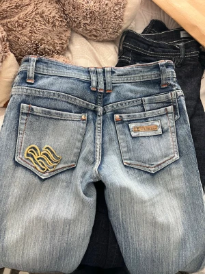 Lågmidjade utsvängda vintage jeans - Säljer ett par snygga ljusblå jeans vintage  med klassisk femficksdesign, orangea sömmar och broderad logga på bakfickan. Jeansen har en något tvättad look och raka ben. Perfekta för en avslappnad stil.