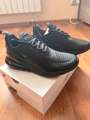 Nike Air Max 270 Triple Black - Nike Air Max 270 Triple Black sneakers med helsvart mesh- och syntetöverdel, stora Air Max-dämpningen i hälen och diskreta svarta swoosh-loggor. Skorna har en sportig siluett, snörning och dragflikar både fram och bak.