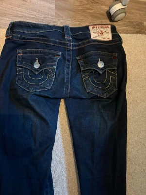 True Religion utsvängda jeans mörkblå - Mörkblå jeans från True Religion med utsvängda ben och snygga detaljerade bakfickor med lock och silverfärgade knappar. Klassisk femficksmodell i denim med kontrastsömmar och läderpatch bak i midjan. Köpta nya för 1 299kr. Varan är i fint skick och är slutsåld på Nelly just nu. De är i storlek 23 i joey modellen med låg midja.