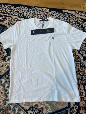 Vit t-shirt från Polo Ralph Lauren - Klassisk vit t-shirt från Polo Ralph Lauren med rund halsringning och ikoniska broderade loggan på bröstet. Tillverkad i mjuk bomull och har en normal passform. Perfekt basplagg för en clean och stilren look.
