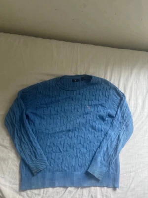 Blå kabelstickad tröja från Gant - Snygg blå kabelstickad tröja från Gant med rund halsringning och diskret broderad logga på bröstet. Tröjan har långa ärmar och ribbade muddar vid ärmslut och nederkant. Perfekt för en stilren och klassisk look.