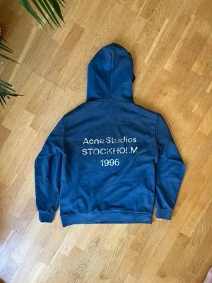  Acne Studios Hoodie  - Snygg blå hoodie från Acne Studios med tryck på ryggen där det står 'Acne Studios STOCKHOLM 1996'. Klassisk passform med huva och ribbade muddar vid ärmslut och nederkant. Perfekt för en chill och trendig look.