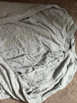 Ljusgrå cardigan från 157 - Ljusgrå cardigan med knappar framtill från 157. Tröjan har ribbstickade muddar vid halsringning och ärmslut. Tillverkad i ett mjukt och tunt material, perfekt för lager-på-lager. Enkel och stilren design som passar till det mesta.