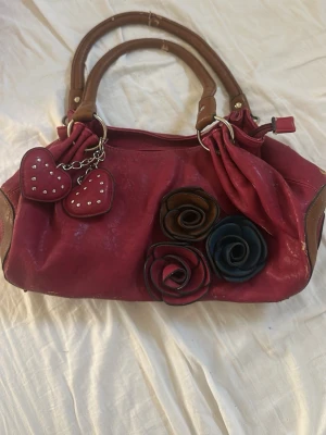 Röd handväska med blommor och hjärtan - Cool röd handväska med bruna handtag och dekorativa blommor i rött, brunt och marinblått på framsidan. Väskan har två hjärtformade nyckelringar med nitar och detaljer i brunt läder. Perfekt för dig som gillar unika accessoarer.