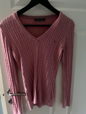 Rosa kabelstickad tröja Tommy Hilfiger - Säljer en rosa kabelstickad tröja från Tommy Hilfiger med V-ringning och lång ärm. Tröjan har en liten logga på bröstet och ribbade muddar vid ärmslut och nederkant. Perfekt för dig som gillar klassisk och stilren design.