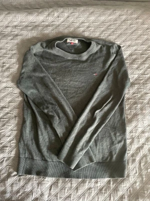 Grå tröja från Tommy Hilfiger - Snygg grå tröja från Tommy Hilfiger med rund halsringning och diskret logga på bröstet. Tröjan har långa ärmar och ribbade muddar vid ärmslut och nederkant. Perfekt för en stilren och enkel look.