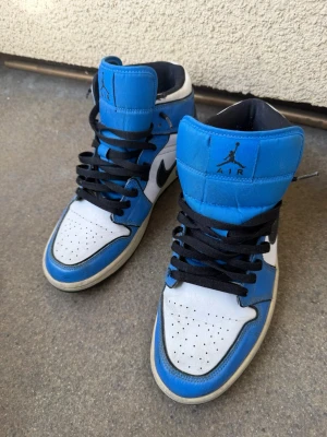 Nike Air Jordan 1 Retro blå/vit - Säljer ett par Nike Air Jordan 1 Retro sneakers i blått, vitt och svart. Klassisk high-top modell med svart snörning och ikonisk Jumpman-logga på plösen. Ovandel i skinn och syntet, perfekt för dig som gillar streetwear.