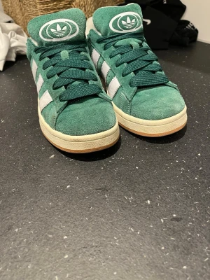 Gröna Adidas sneakers i mocka - Snygga gröna Adidas sneakers i mocka med klassiska vita ränder på sidorna och retro Adidas-logga på plösen. Skorna har gröna skosnören och en vit sula med gummidetaljer. Perfekta för dig som gillar streetwear och vill sticka ut.