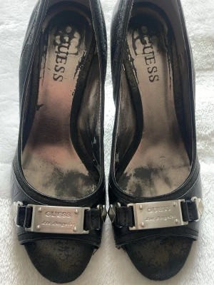 Vintage Guess klackskor  - Snygga vintage svarta klackskor från Guess med peep toe och hög klack. Skorna har en mix av läder och textil, samt en metallplatta framtill med Guess-logga och nitar som cool detalj. Perfekta för dig som vill ha en edgy och trendig look. Kan mötas upp i Stockholm!