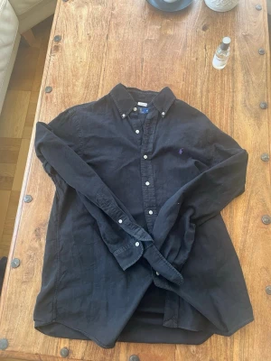 Svart skjorta från Polo Ralph Lauren - Säljer en svart skjorta från Polo Ralph Lauren med klassisk button-down krage och vita knappar. Skjortan har lång ärm och är tillverkad i bomull. Diskret lila logga broderad på bröstet. Perfekt för en stilren look. Passar runt 180