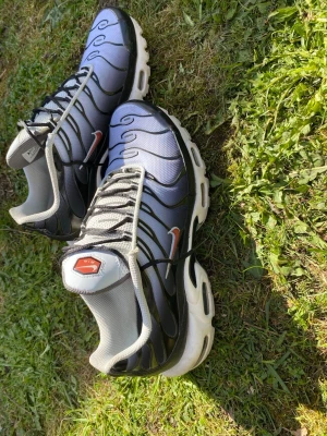 Nike Air Max Plus TN svart/vit/Blåa - Nike Air Max Plus TN sneakers i svart och vitt med ikoniska vågmönster och röd Swoosh på sidan. Ovandelen är i mesh och syntet, med vita och svarta detaljer samt synlig Air-dämpning i sulan. Klassisk TN-logga på plösen och robust yttersula. Använda några fåtal gånger ser helt nya ut