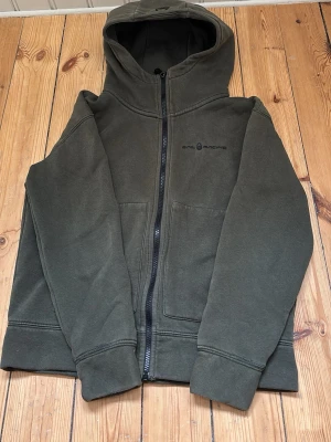 Mörkgrön hoodie från Sail racing  - Mörkgrön hoodie med dragkedja från Sail racing. Klassisk modell med huva och två stora fickor framtill. Tillverkad i mjukt och bekvämt material, perfekt för chill dagar. Diskret logga på bröstet och breda muddar vid ärmslut och nederkant.