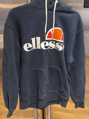 Mörkblå Ellesse hoodie med logga - Snygg mörkblå hoodie från Ellesse med stor vit och orange logga på bröstet. Tröjan har huva med vita snören och en praktisk magficka. Perfekt för dig som gillar sportig och avslappnad stil. Materialet är mjukt och bekvämt, troligen bomullsblandning.