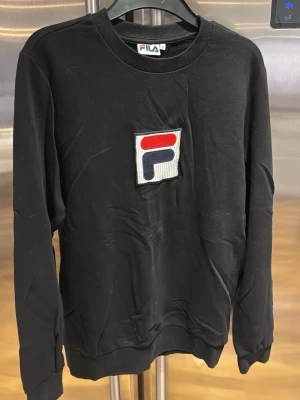 Svart sweatshirt från FILA - Cool svart sweatshirt från FILA med stort broderat logotypmärke på bröstet i rött, blått och vitt. Tröjan har rund halsringning och ribbade muddar vid ärmslut och nederkant. Perfekt för en avslappnad och sportig look.