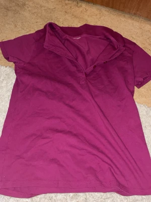 Mörkrosa pikétröja med krage - En enkel och stilren mörkrosa pikétröja med korta ärmar och klassisk krage. Tröjan har knappar vid halsen och är gjord i mjukt bomullstyg. Perfekt för en avslappnad och snygg look.