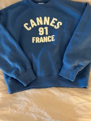 Blå sweatshirt med Cannes-tryck - Cool blå sweatshirt med vitt texttryck 'Cannes 91 France' på bröstet. Tröjan har rund hals, ribbade muddar och är tillverkad i mjukt material som känns skönt mot huden. Perfekt för dig som gillar sportiga och avslappnade looks.