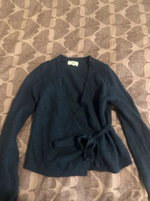 Mörkblå omlottkofta med knytband - Mysig mörkblå omlottkofta med långa ärmar och knytband i midjan. Koftan har en mjuk och stickad känsla, perfekt för lager-på-lager och chill dagar. Enkel och stilren design som är lätt att matcha med jeans eller kjol.