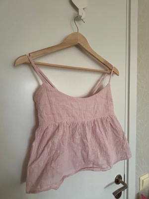 Ljusrosa linnetopp med knyt i ryggen från Nelly - Supersöt ljusrosa linnetopp med tunna axelband och volang nedtill. Toppen har en snygg knytning i ryggen som ger en extra detalj. 