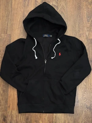Svart hoodie från Polo Ralph Lauren - (Storlek L men sitter som M!) Svart hoodie med dragkedja från Polo Ralph Lauren. Klassisk design med vit dragsko i huvan och röd broderad logga på bröstet. Mjuk bomullsmix och stora fickor framtill. Perfekt för en avslappnad look. 