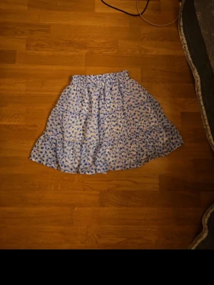 Blommig blå kortkjol med volang - Söt kortkjol med blå blommigt mönster på vit bakgrund. Kjolen har volangdetaljer och elastisk midja, perfekt för en avslappnad och trendig look. Materialet känns lätt och luftigt, idealiskt för varmare dagar.