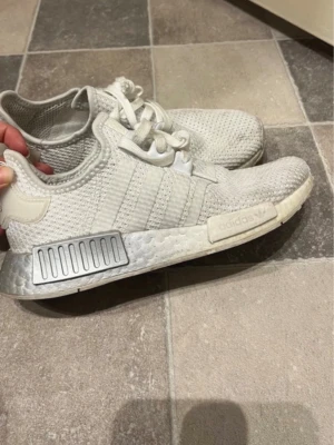 Adidas NMD R1 vita sneakers - Snygga vita Adidas NMD R1 sneakers med stickad mesh-överdel och klassiska tre ränder på sidan. Skorna har Boost-sula och detaljer i silver och vitt. Perfekt för dig som gillar streetstyle och komfort.