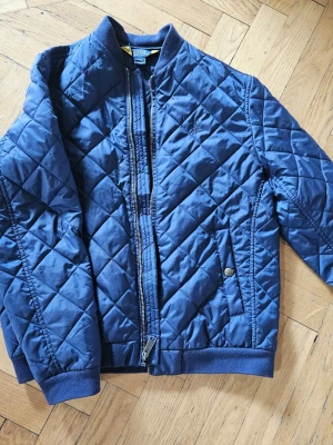 Ralph Lauren jacka stl 14-16 - Snygg marinblå pufferjacka från Polo Ralph Lauren i storlek L (14-16). Jackan har quiltat mönster, ribbade muddar och krage samt dragkedja framtill. Två fickor med knappdetalj och diskret logga på bröstet. Perfekt för kalla dagar.