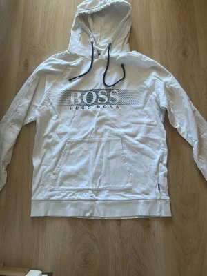 Vit hoodie från Hugo Boss - Snygg vit hoodie från Hugo Boss med svart logga och text på bröstet. Hoodien har huva med svarta dragsnören och en stor magficka framtill. Tillverkad i mjukt bomullsmaterial och har ribbade muddar vid ärmslut och nederkant.