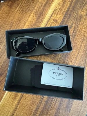 Svarta solglasögon från Prada - Snygga svarta solglasögon från Prada med klassisk och stilren design. Glasögonen har en bred båge i svart syntetmaterial och kommer med originalbox och äkthetskort. Perfekt för dig som vill ha en exklusiv accessoar med ikonisk look. OBS de är lite små i storlek hör av er vid frågor :)