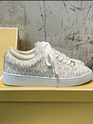 Vita MK sneakers med logomönster - Snygga vita sneakers från Michael Kors med klassiskt MK-logomönster över hela skon. Skorna har vita snören med guldfärgade detaljer och en vit gummisula. Perfekt för dig som vill ha en trendig och stilren look.