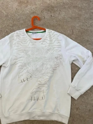 Vit sweatshirt från Kenzo med tiger - Cool vit sweatshirt från Kenzo med broderad tiger och logotyp på framsidan. Tröjan har rund hals, ribbade muddar och detaljer i form av broderade linjer både fram och bak. Perfekt för dig som gillar streetstyle och exklusiva märken.