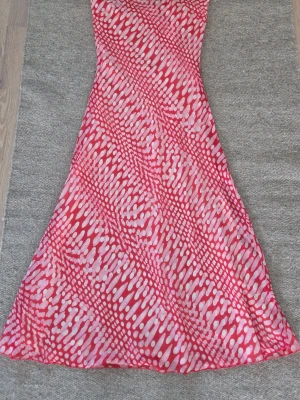Rosa långklänning med prickigt mönster - Cool långklänning i rosa och vitt med ett unikt prickigt mönster. Klänningen har hög krage med knappar och är ärmlös, vilket ger en snygg och modern look. Materialet känns lätt och passar perfekt för varma dagar o ljumma  sommarnätter.