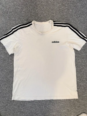 Vit Adidas t-shirt med svarta ränder - Säljer en klassisk vit t-shirt från Adidas med svarta ränder längs axlarna och logga på bröstet. Den är 67cm på längden och 52cm på bräden. Hör av dig frågor och funderingar!🤗