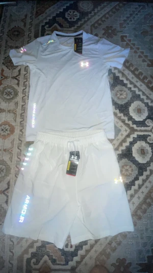 Vitt set från Under Armour helt ny med tag - Säljer ett vitt set från Under Armour med t-shirt och shorts. Båda plaggen har reflekterande loggor och text, och är tillverkade i ett lätt och sportigt material som passar perfekt för träning eller chill. T-shirten har rund hals och korta ärmar, shortsen har elastisk midja med snörning.