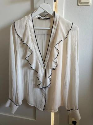 Vit blus med volang och svarta detaljer - Säljer en superfin vit blus med volang längs v-ringningen och svarta kantdetaljer. Blusen har ett skönt material som passar perfekt till en härlig sommarkväll eller bara till vardags. Hör gärna av dig vid frågor!💗