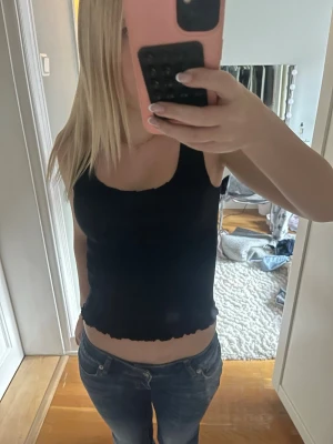 Svart ribbad linnetopp - Säljer en svart ribbad linnetopp med vågig kant nertill. Toppen är ärmlös och har en tight passform som sitter snyggt på kroppen. Perfekt att matcha med jeans eller kjol för en enkel och stilren look.