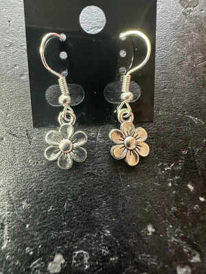 Silvriga blommiga örhängen - Säljer ett par silvriga örhängen med hängande blommor. Örhängena har en enkel krok och blomman har fina detaljerade kronblad. Perfekta för dig som gillar söta och stilrena accessoarer.
